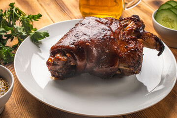 Baked pork knuckle on a white plate on a wooden background or table with bread, fresh cucumbers, parsley and mustard and a mug of beer. Traditional Bavarian dish.