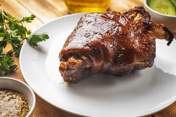 Baked pork knuckle on a white plate on a wooden background or table with bread, fresh cucumbers, parsley and mustard and a mug of beer. Traditional Bavarian dish.