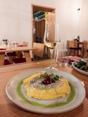 Delicious dish, white plate with rabbit stew and mashed potatoes. The dish is standing on wooden table in a restaurant. Background of restaurant table, drinking glasses. Light green sauce on the plate