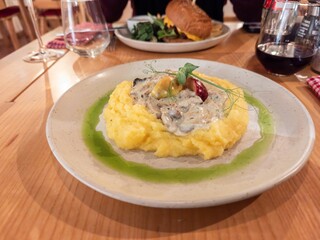 Delicious dish, white plate with rabbit stew and mashed potatoes. The dish is standing on wooden table in a restaurant. Background of restaurant table, drinking glasses. Light green sauce on the plate