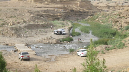 Several cars by a river in a scenic landscape, showing people enjoying nature.