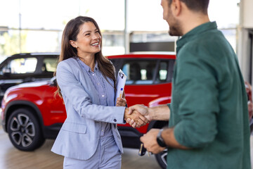 Beautiful young woman is talking to handsome car dealership worker while choosing a car in...