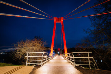 Symmetrical view of an illuminated pedestrian bridge with vibrant red lighting, set against a deep night sky, with cables converging toward the structure..