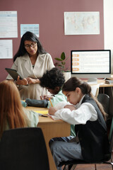 Fototapeta premium Group of clever intercultural schoolkids sitting by table in classroom and writing down English grammar rules in copybooks