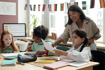 Happy young teacher of English grammar passing paper with test to cute schoolgirl sitting by desk...