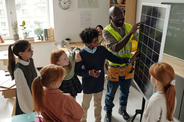 Cute primary schoolboy and professional engineer touching solar panel during presentation of...