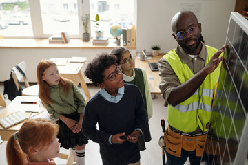 Confident technician touching model of solar panel while standing among schoolkids during...