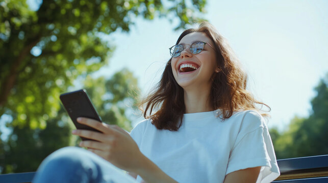A young woman in a park, sitting on a bench, holding her smartphone and laughing out loud as she enjoys the fresh air and sunny weather.