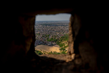  Jaipur framed through a small stone window, emphasizing the contrast between the ancient architecture and the sprawling modern city. The scene highlights greenery, sand, and dense urban sprawl