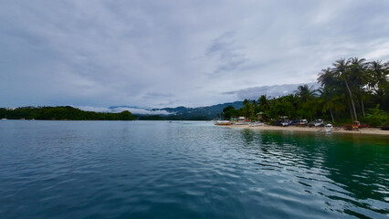 Fishing village on the shore of a tropical island. View of the strait between tropical islands, hilly coastline and a settlement on the seashore, sandy beach and palm trees.
