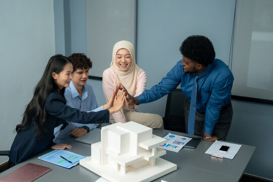 Diverse group of professionals high-fiving over a table with architectural models and documents, celebrating successful collaboration in an office.