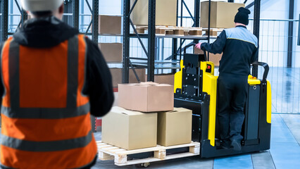 Teamwork of warehouse employees. Electric forklift for moving pallets. Foreman supervises work of warehouse employees. Warehouse manager stands with back to camera. Workers inside storage building