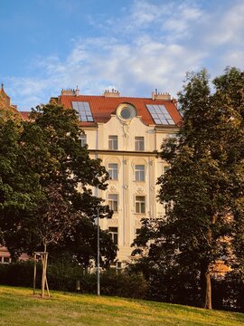 Golden hour view of a historic building in Prague's Vinohrady district, framed by lush green park trees and a vibrant blue sky. The intricate facade glows warmly in the evening sunlight.