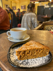 Cozy cafe scene with delicious slice of cake and cup of coffee or tea on table over blurred people in a warm ambient setting. 