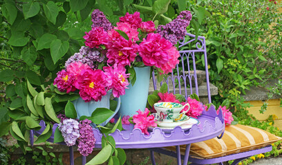 Red camellias and lilac floral still life in blue vases on a purple metal table with cup and saucer outdoors in a garden
