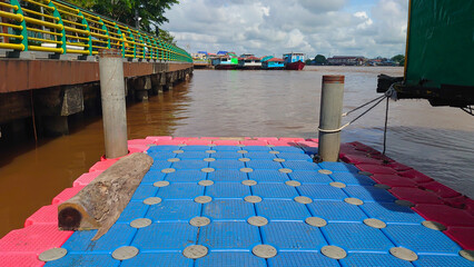 A vibrant floating dock with blue and red panels on a calm river, surrounded by boats and a railing walkway. The tranquil scene captures the essence of waterfront life.