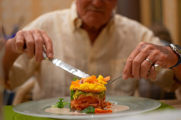 Elderly man at restaurant eats healthy gourmet salad plate consisting of fruit, vegetables and salmon, papaya, avocado, mango