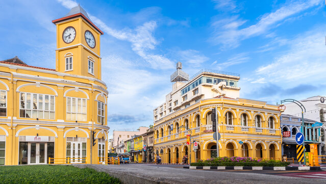 The historic Sino-Portuguese clock tower and Chartered Bank building in Phuket Old Town frame a bustling intersection, with a distinctive blue local bus passing under a clear blue sky