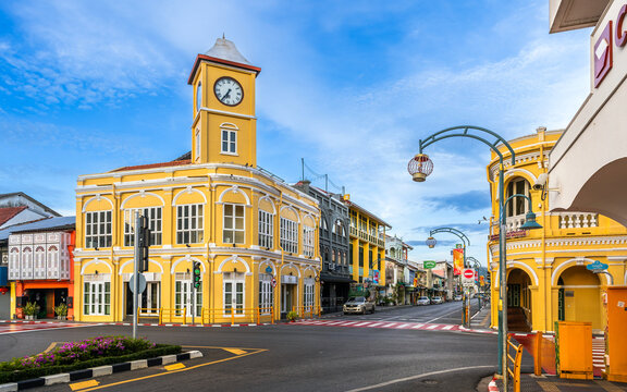 The iconic Sino-Portuguese clock tower in Phuket Old Town stands at a vibrant intersection surrounded by colorful colonial-style buildings under a bright blue sky