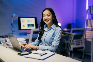 Young asian businesswoman smiling and working late using laptop computer in office at night with computer and documents on desk, she is focused on her work