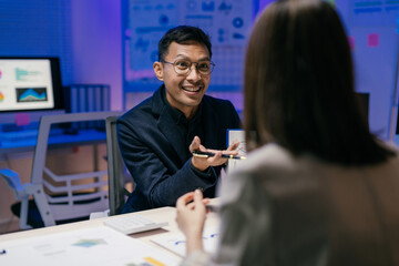 Businessman is holding a pen and explaining a business strategy to his colleague in a modern office at night, surrounded by computers and documents