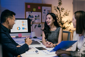 Three asian businesspeople are discussing a financial report in an office at night, using a computer and printed documents, working overtime