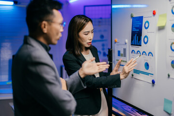 Businesswoman pointing at charts and graphs on a whiteboard while explaining business strategy to a businessman in a modern office with blue lighting
