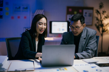 Two asian businesspeople are working together on a laptop in a modern office at night, collaborating on a project with documents and charts spread out on the desk