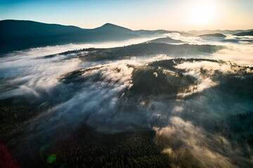 Stunning aerial view of sunrise over mountainous landscape, with thick fog blanketing valleys and hills. Sun's golden rays pierce through mist, casting serene glow over the entire scene.