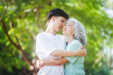 Fototapeta premium Young son and his mature mom asian people share loving hug in beautiful park, surrounded by nature. Their happiness radiates as they bond outdoors, celebrating family love and togetherness.