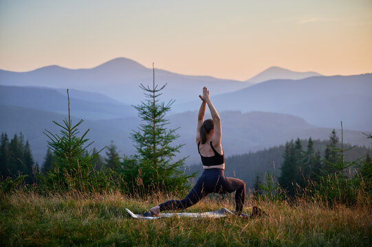 Woman practicing yoga and meditating outdoors in the mountains in a serene, natural setting. Female performing yoga pose on mat, with backdrop of beautiful mountain landscape at sunrise or sunset.