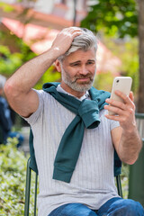 Fototapeta premium Bearded mature man using smartphone typing browsing, loses becoming surprised sudden lottery results, bad news, fortune loss, fail. Tourist guy standing in urban city street outdoors. Town lifestyles.