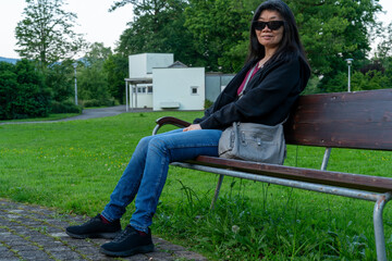 Asian middle aged woman with sun glasses sitting on a bench in the park, having a rest. Green nature with white building in the background.