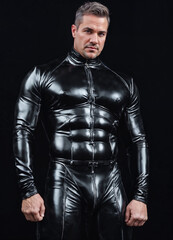 handsome man in a black leather suit poses confidently in a dark studio. The lighting highlights his muscular physique and the sleek, shiny fabric.