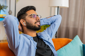 Happy relaxed young Indian man resting on couch with wireless earphones listening to ambient calming tranquil music from online playlist. Smiling Hispanic guy resting with hands behind head in room.