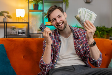 Successful rich man holding smartphone and waving dollar bill money fan sitting on sofa at home. Happy excited Caucasian guy winning online casino lottery game planning vacation calculating budget