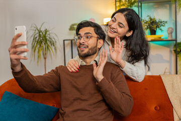 Diverse happy Indian couple waving hello while talking on smartphone video call on sofa in room at home. Cheerful girlfriend and boyfriend in casual clothes recording vlog on cellphone in apartment.