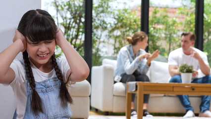Stressed and unhappy young girl huddle in corner, cover her ears blocking sound of her parent arguing in background. Domestic violence at home and traumatic childhood develop to depression. Synchronos