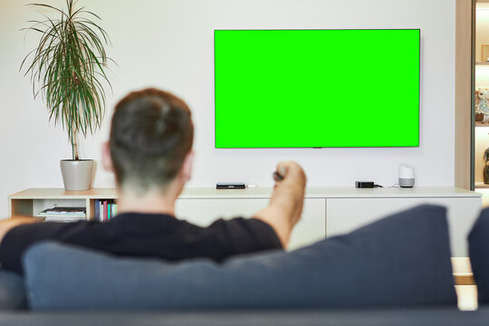 Rear view. The guy turns on the plasma flat-screen TV by pointing the remote at the green screen. The man is out of focus, watching TV while relaxing in his living room. Layout for the design. - Powered by Adobe