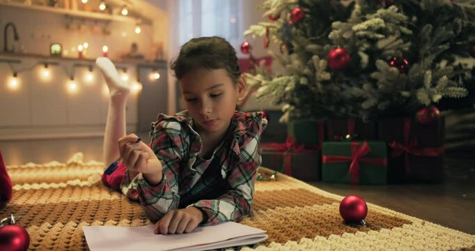 Merry Christmas. Cute kid girl writing letter to Santa on Christmas Eve lying in decorated room. Xmas celebration holiday