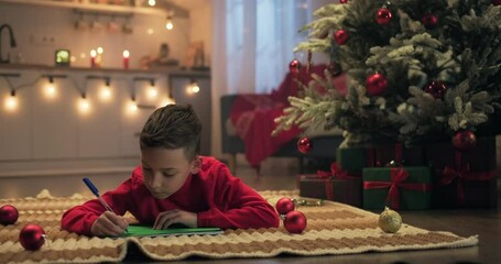 Merry Christmas. Cute boy writing letter to Santa on Christmas Eve lying in decorated room. Dreaming kid thinking of wish saying oh. Xmas celebration holiday