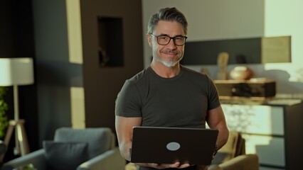 Happy mid adult man in his 50s working on laptop computer standing in modern living room. Portrait of businessman managing business online in home office, smiling.