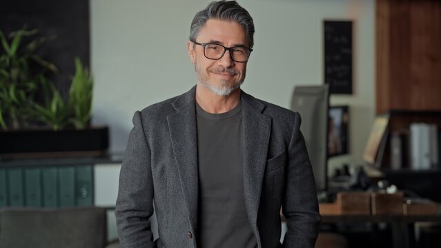 Confident mature businessman standing in office. Entrepreneur managing business online Portrait of happy mid adult, middle aged man in glasess with gray hair, smiling.