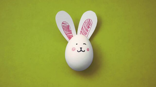 4K video. White chicken egg with cute rabbit face and bunny ears on yellow background. Top view. Gift hunt. Magic frame-by-frame stop motion animation for Happy Happy Easter. Greeting card for holiday