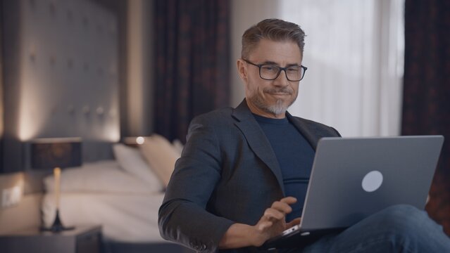 Professional man in business casual using laptop in hotel room. Businessman working at home with computer, smiling. Happy mid adult male with content smile. Entrepreneur managing business online.
