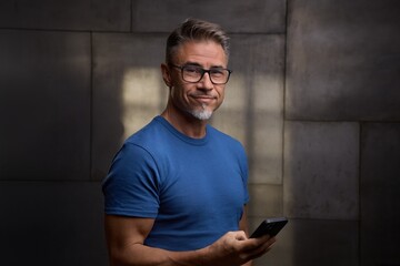 Confident mature man standing against dark industrial wall in office, using mobile phone. Portrait...