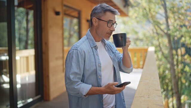 Mature man drinking first cup of coffee at morning while using smartphone to check news and message on the balcony of a modern wooden chalet, surrounded by trees and a peaceful natural forrest. - Powered by Adobe