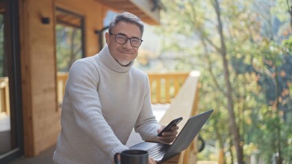 Mature businessman using laptop and smartphone on wooden terrace of mountain chalet, remote working...