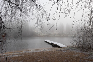 A foggy morning by the lake with a wooden dock surrounded by trees and distant houses in a tranquil...