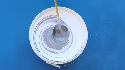 Bucket of white wall paint that is mixed with mixer with blue and black paint to get gray color with light blue tint.